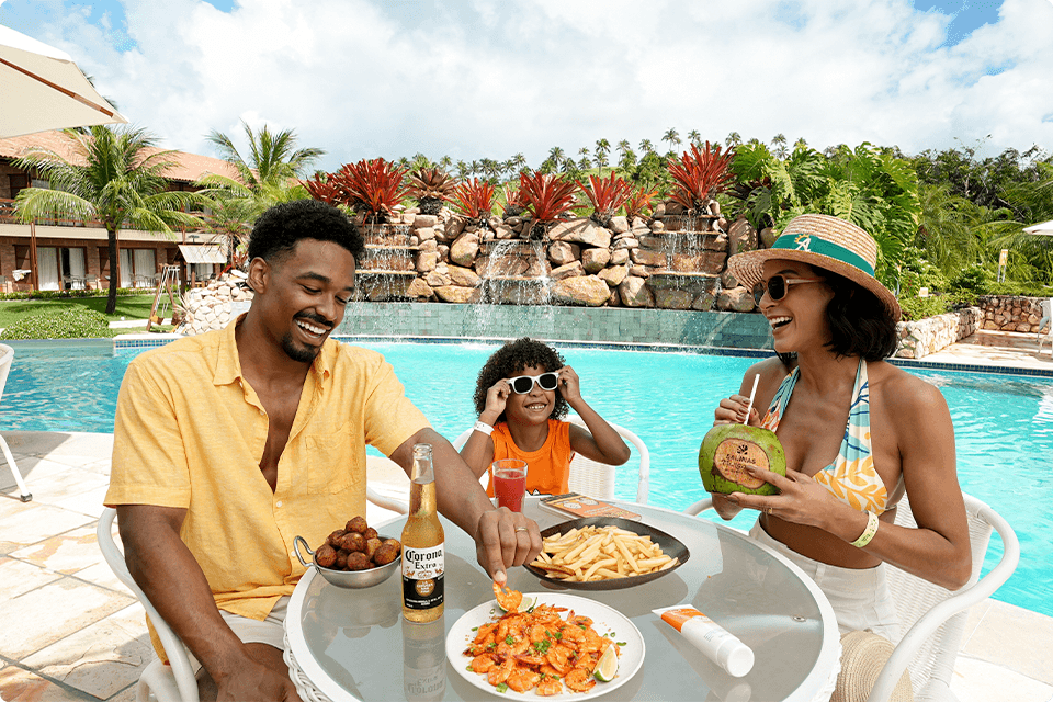 Família com prato de petiscos com camarão, bolinha de queijo e batata frita, as bebidas sendo cervejas, suco e água de coco sobre mesa à beira da piscina do Salinas Maragogi com cachoeira artificial atrás rodeando de plantas.