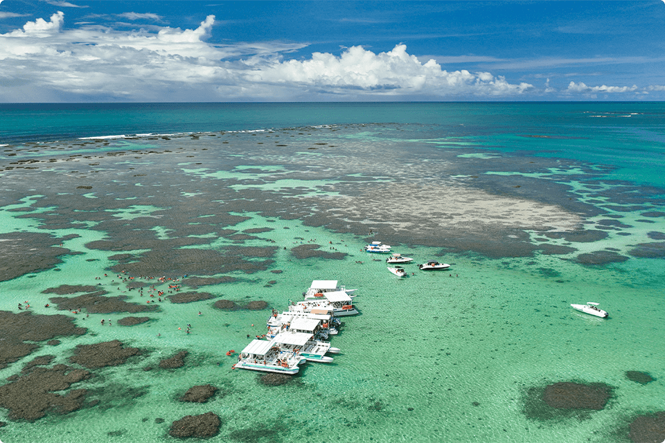 Vários catamarãs nas piscinas naturais da praia de Galés em Maragogi. O céu está com algumas nuvens, é possível enxergar os recifes de corais e há muitas famílias na água cristalina.