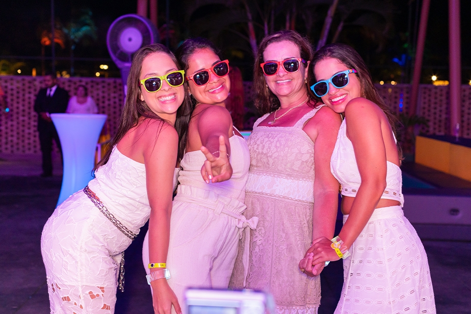 Quatro mulheres usando roupas brancas em comemoração ao ano novo e
 óculos escuros coloridos posam pra a foto, sorrindo, na festa do
 Réveillon no Salinas Maragogi.