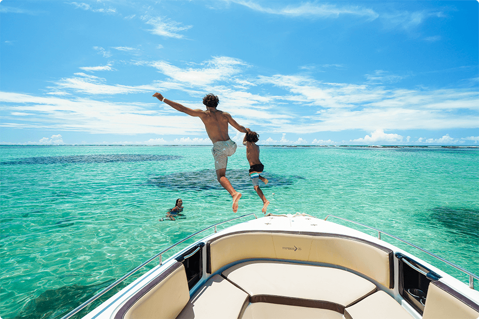 Vista da praia de Galés, com uma lancha em águas cristalinas e
piscinas naturais, pai e filho estão pulando no mar e mãe já está na água proporcionando um cenário paradisíaco.