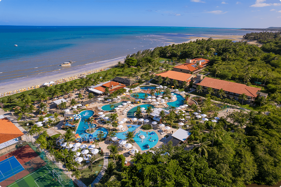 Vista aérea do Salinas Maragogi All Inclusive Resort, mostrando
a área de piscinas, guarda-sóis brancos e estrutura do resort, cercado por vegetação e está à beira-mar.