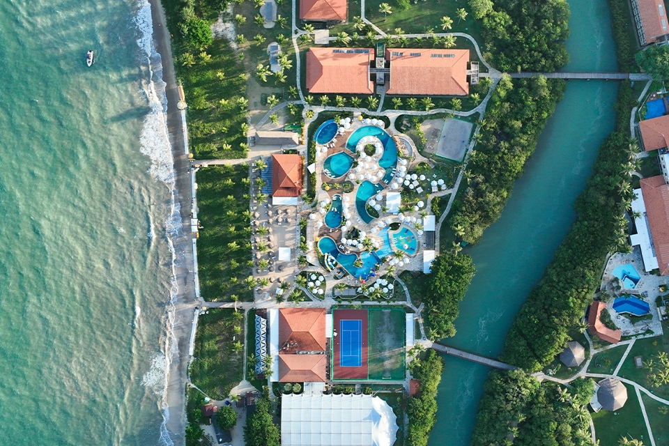 Vista aérea do Salinas Maragogi All Inclusive Resort, mostrando
a área das piscinas e algumas acomodações que são cortadas pelo Rio Maragogi. Todo o resort é cercado por vegetação tropical, palmeiras e está à beira-mar.