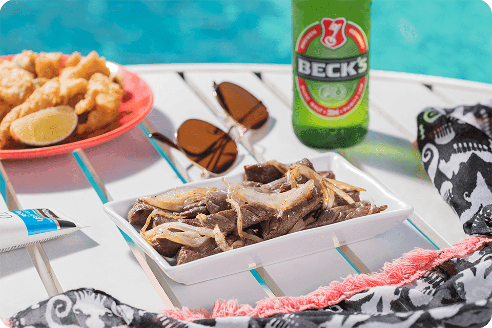 Cerveja Becks e pratos com iscas de carne bovida acebolada e camarão empanado sobre mesa à beira da piscina no Salinas Maragogi.