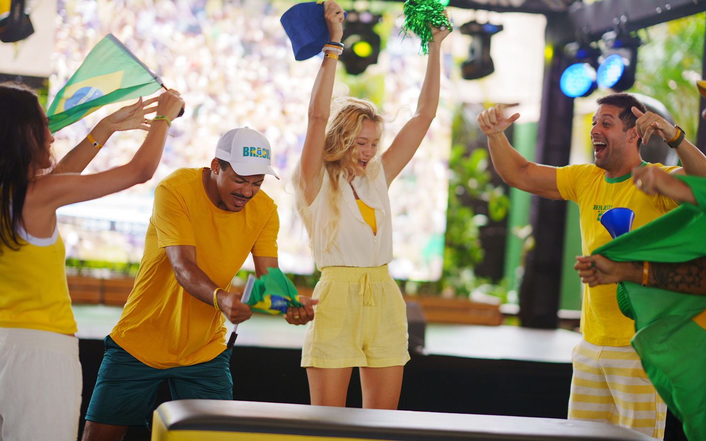  Grupo de amigos comemorando o jogo do Brasil na copa com roupas amarelas e verdes, com acessórios como a bandeira do Brasil, chapéus. E há um palco ao fundo com telão pra transmissão.
