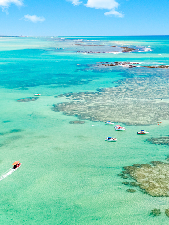 Vista aérea do mar cristalino da Praia de Barra Grande com tons azul-turquesa e catamarã navegando em meio às piscinas naturais.