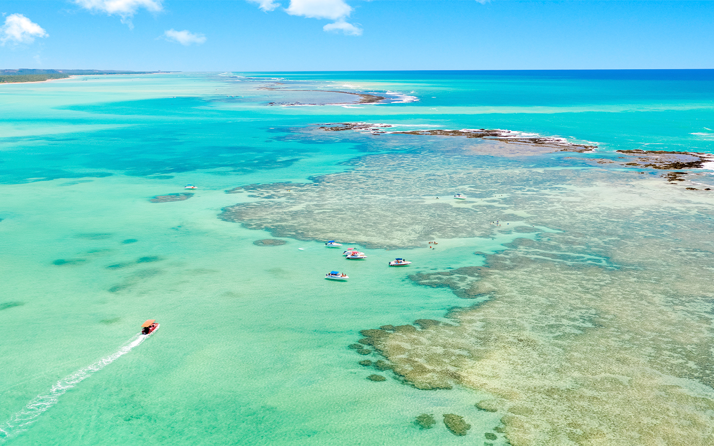 Vista aérea do mar cristalino da Praia de Barra Grande com tons azul-turquesa e catamarã navegando em meio às piscinas naturais.