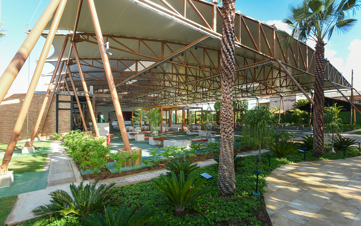 Ampla estrutura do Espaço Cabanos coberta com vários bancos e cadeiras, paisagismo tropical verde.