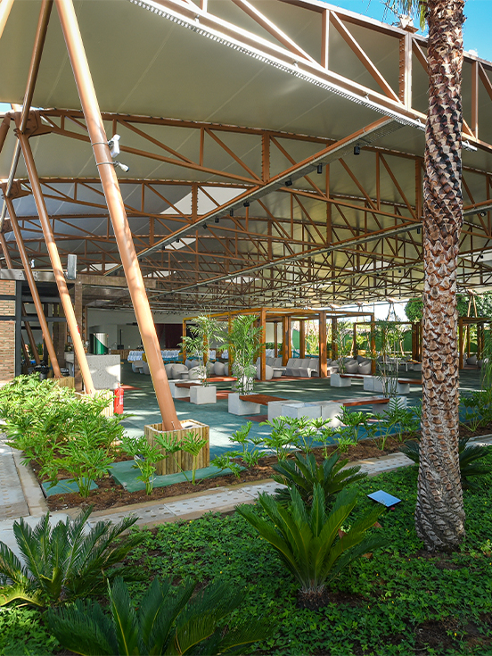 Ampla estrutura do Espaço Cabanos coberta com vários bancos e cadeiras, paisagismo tropical verde.