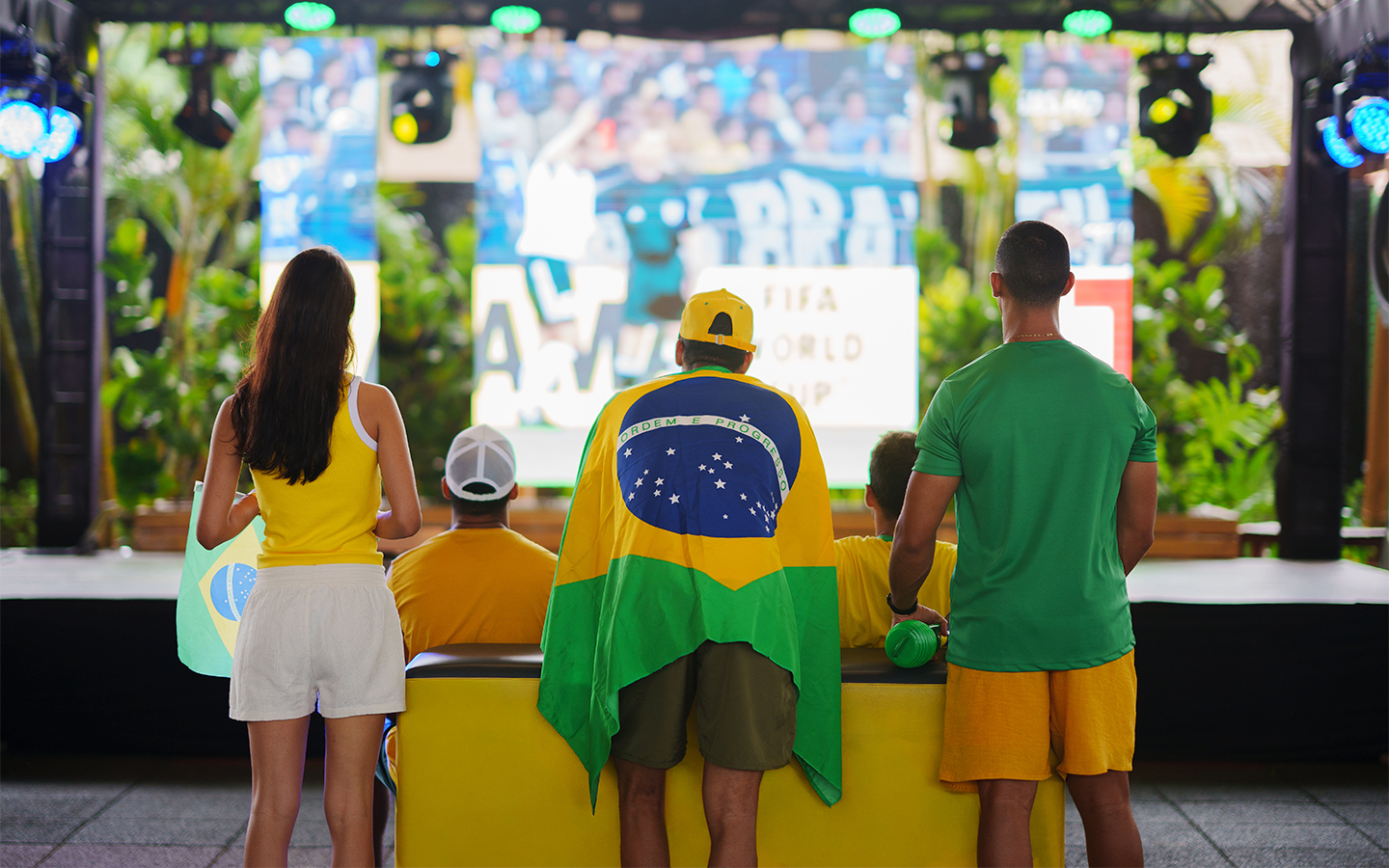 Grupo de pessoas vestidas com as cores da bandeira e camisas amarela e verde, de costas, assistindo à transmissão em telão gigante no Espaço Cabanos.