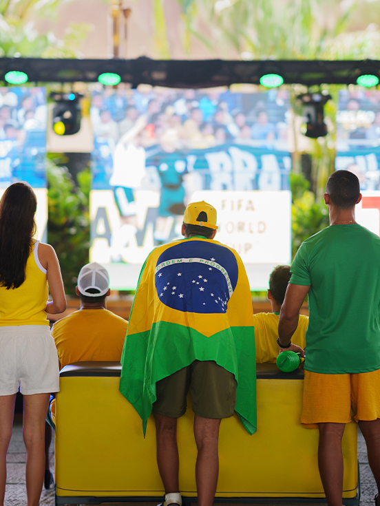 Viva a emoção da torcida pelo hexa Grupo de pessoas vestidas com as cores da bandeira e camisas amarela e verde, de costas, assistindo à transmissão em telão gigante no Espaço Cabanos.