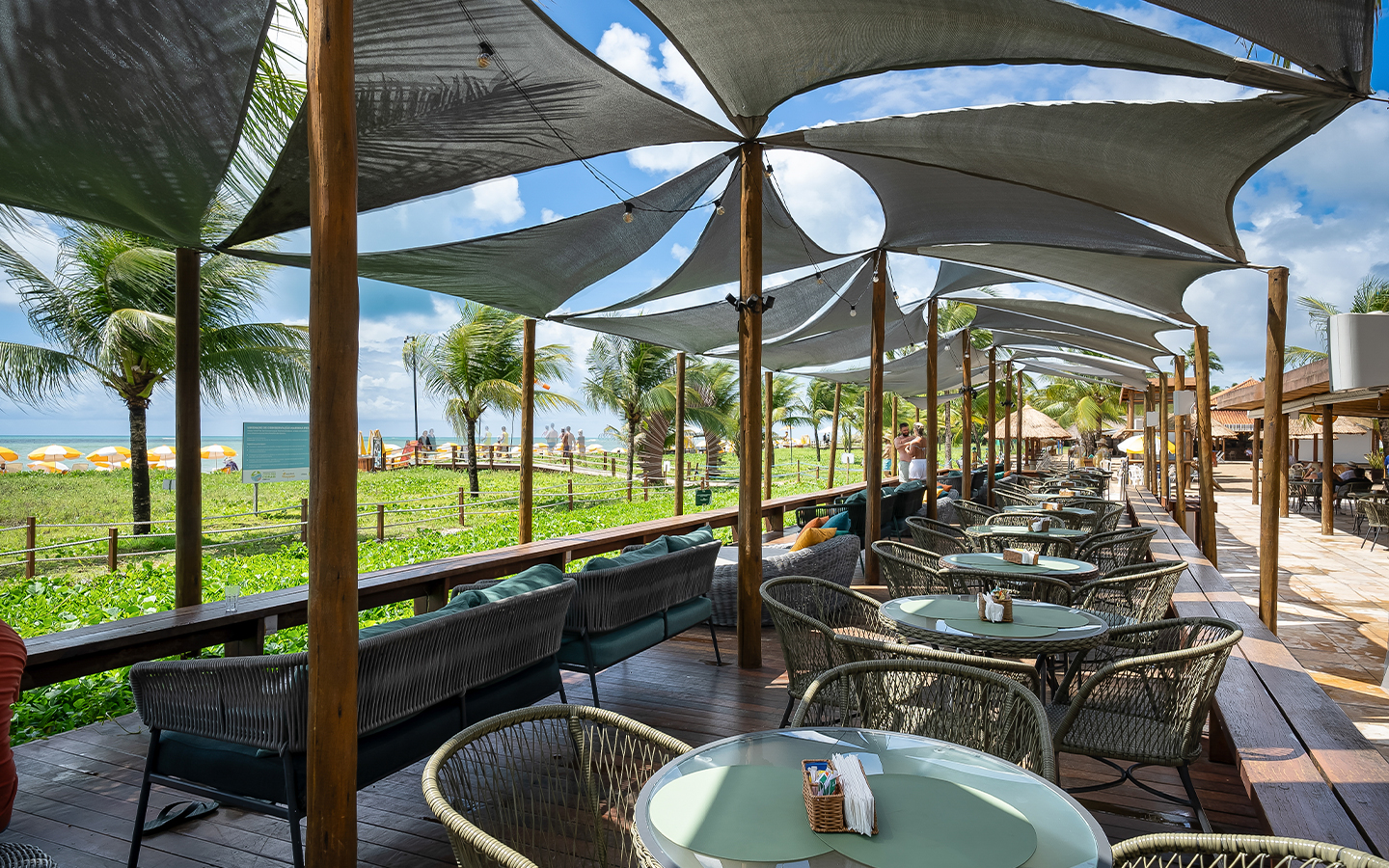 Área externa pra aproveitar o All Inclusive Área externa do Salinas Maragogi e lounge à beira-mar com mesas redondas, cadeiras de corda náutica e cobertura tensionada cinza. O dia está com algumas nuvens e ao fundo há vegetação verde.