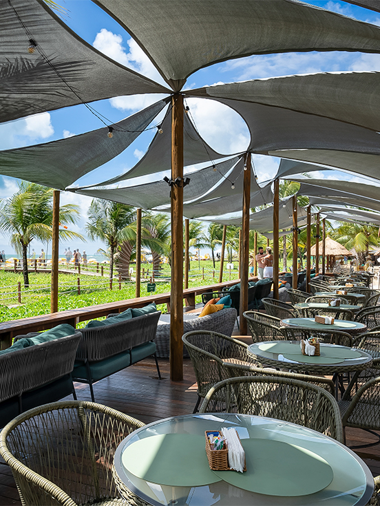 Área externa pra aproveitar o All Inclusive Área externa do Salinas Maragogi e lounge à beira-mar com mesas redondas, cadeiras de corda náutica e cobertura tensionada cinza. O dia está com algumas nuvens e ao fundo há vegetação verde.