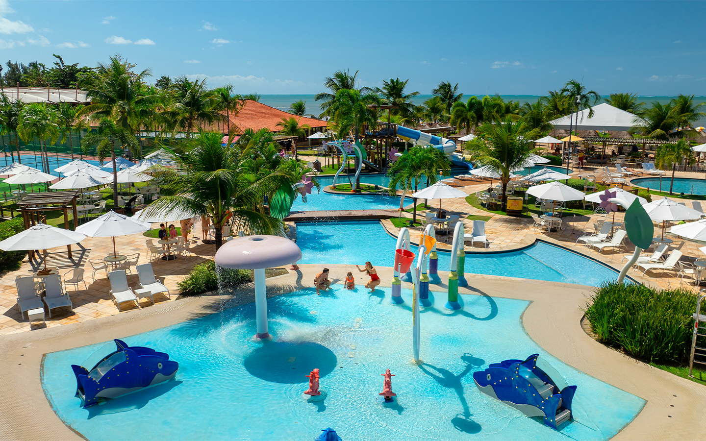 Área externa do Salinas Maragogi com complexo de piscinas, espreguiçadeiras, guarda-sóis e família brincando na piscina infantil. O dia está sem nuvens e ao fundo há vegetação verde.