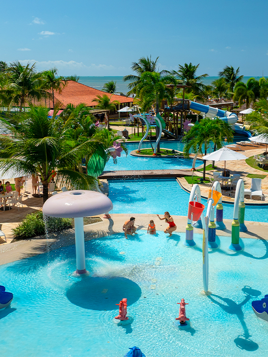 Área externa do Salinas Maragogi com complexo de piscinas, espreguiçadeiras, guarda-sóis e família brincando na piscina infantil. O dia está sem nuvens e ao fundo há vegetação verde.