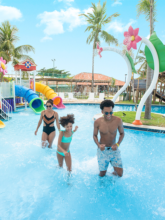Área de lazer do Salinas Maragogi Família com pai, mãe e criança brincando na piscina infantil do Salinas Maragogi, com brinquedos pra crianças, coqueiros, espreguiçadeiras e guarda-sóis ao redor.