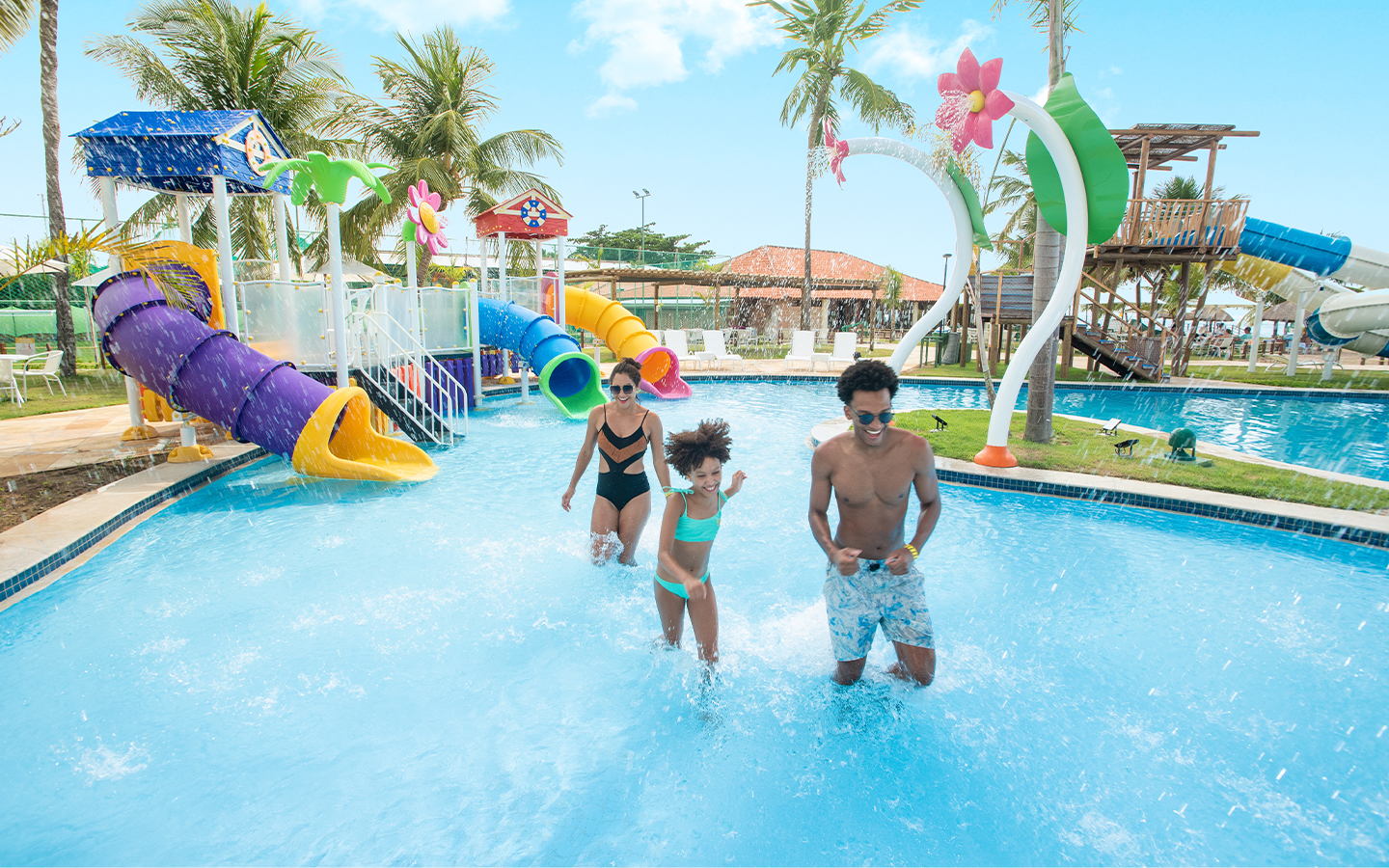 Área de lazer do Salinas Maragogi Família com pai, mãe e criança brincando na piscina infantil do Salinas Maragogi, com brinquedos pra crianças, coqueiros, espreguiçadeiras e guarda-sóis ao redor.