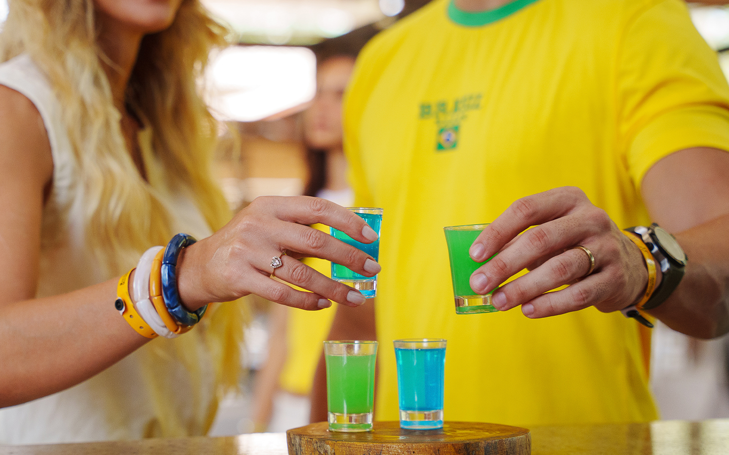 Brinde ao hexa com nossa bebida especial Close-up de mãos segurando copos de shot com bebidas coloridas em azul e verde, com pessoas ao fundo vestindo camisa amarela da seleção.