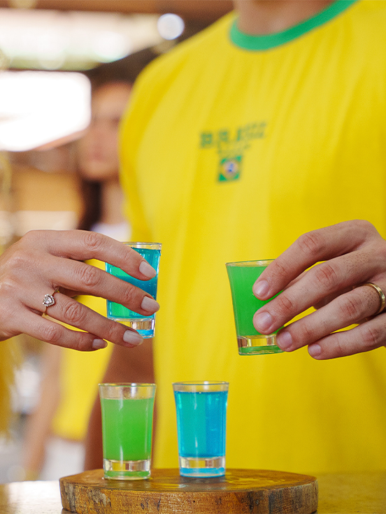 Brinde ao hexa com nossa bebida especial Close-up de mãos segurando copos de shot com bebidas coloridas em azul e verde, com pessoas ao fundo vestindo camisa amarela da seleção.