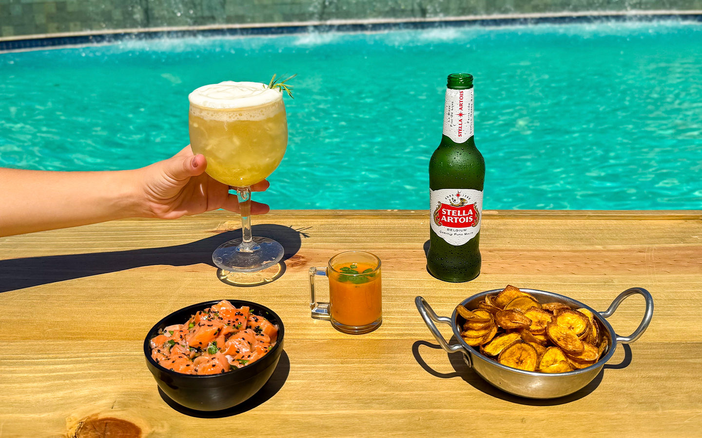All Inclusive pra todos os gostos Mão segurando taça de drink refrescante ao lado de cerveja Stella Artois, bowl de salmão fresco e chips crocantes em mesa de madeira à beira da piscina.