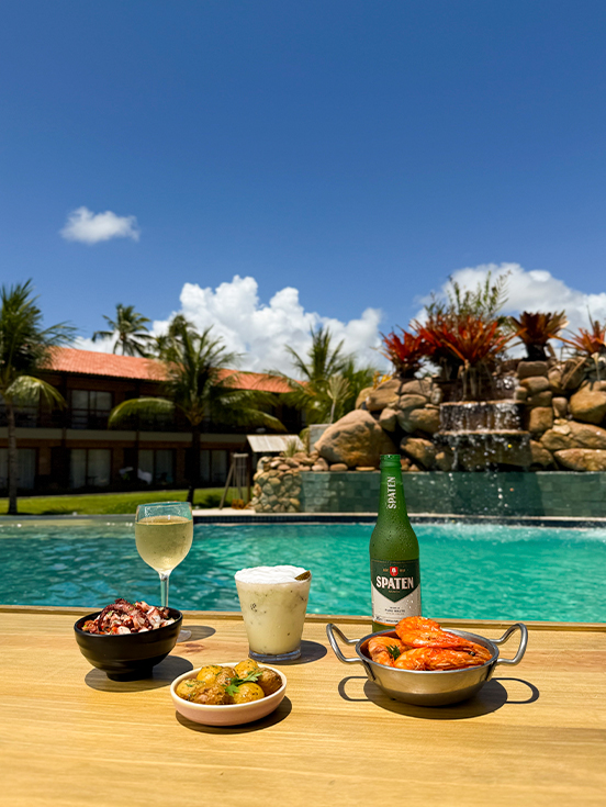 All Inclusive pra todos os gostos Mão segurando taça de drink refrescante ao lado de cerveja Stella Artois, bowl de salmão fresco e chips crocantes em mesa de madeira à beira da piscina.