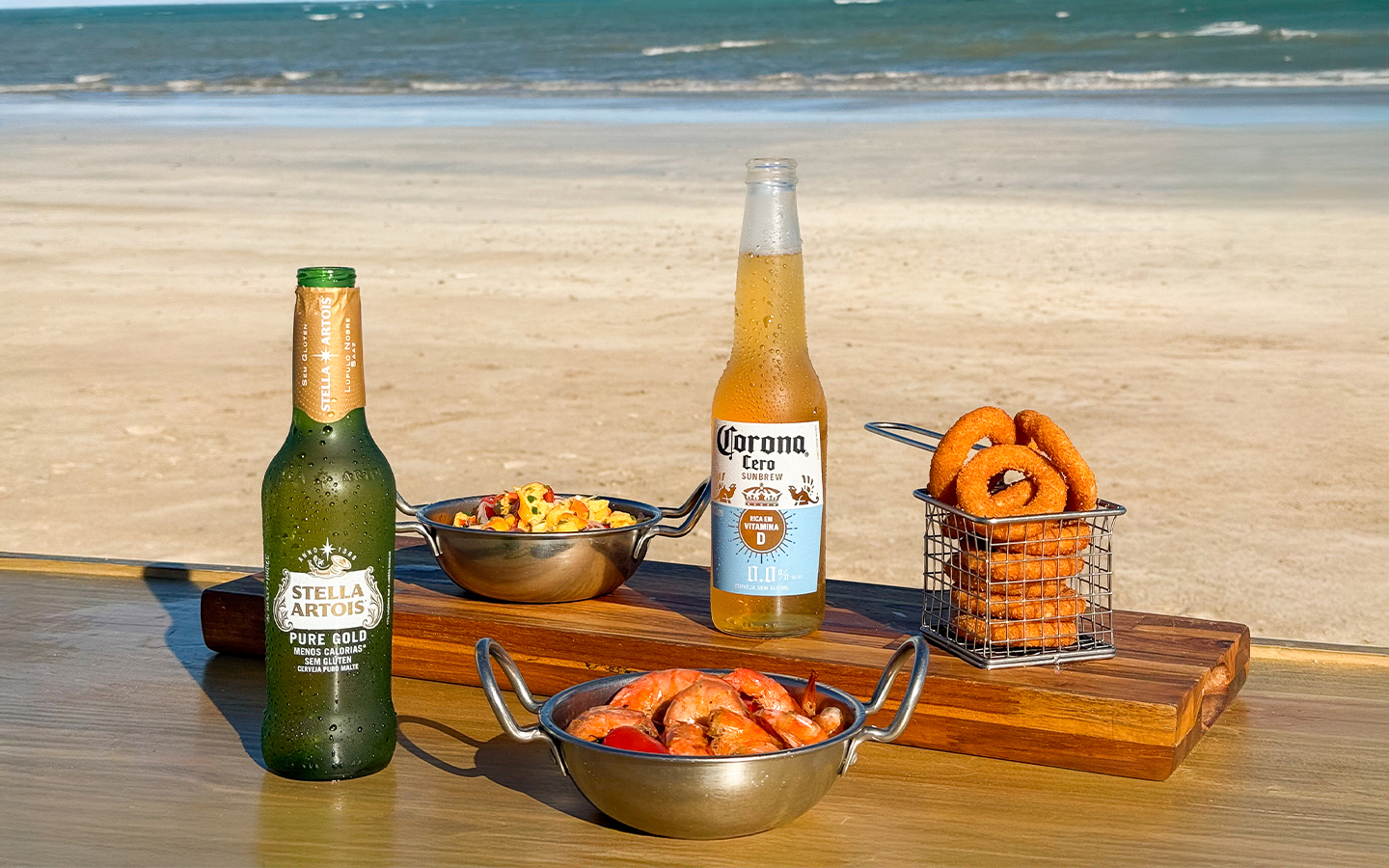 Aproveite nossa oferta de petiscos e bebidas geladas Tábua de madeira com cervejas Stella Artois Pure Gold e Corona, acompanhadas de porções de camarão, lula e onion rings em uma mesa de frente pro mar.