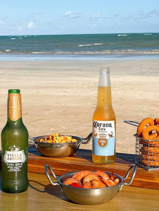 Aproveite nossa oferta de petiscos e bebidas geladas Tábua de madeira com cervejas Stella Artois Pure Gold e Corona, acompanhadas de porções de camarão, lula e onion rings em uma mesa de frente pro mar.