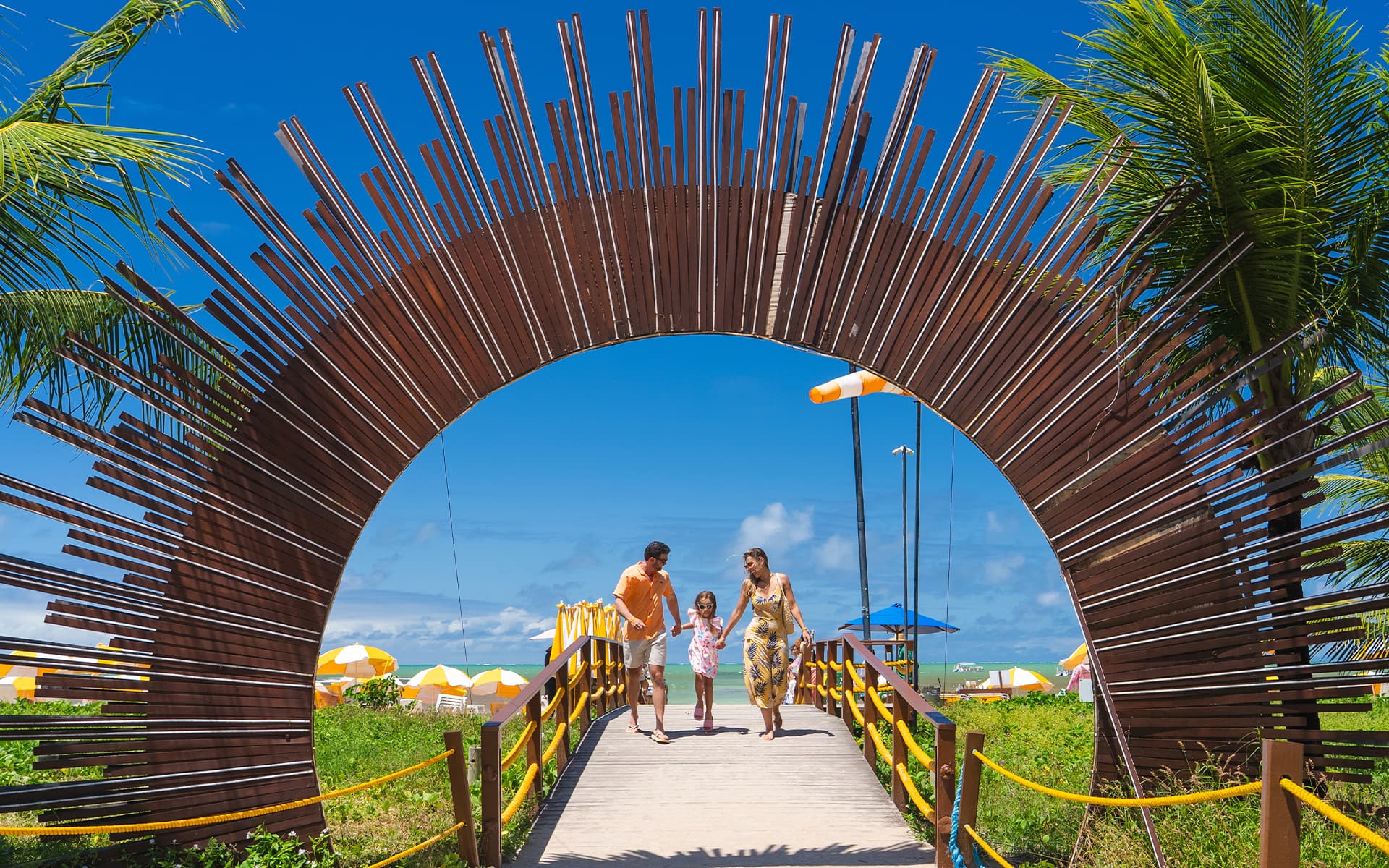 Onde as férias começam Portal do Salinas Maragogi cercado por coqueiros na beira do mar com céu azul.