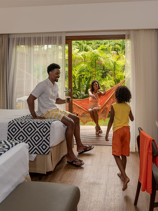 Apartamento Standard Vitória Régia Família de pai sentado na cama, filho em pé e mamãe na rede da varanda do apartamento Standard Vitória Régia no Salinas Maragogi com conforto pra hospedagem.