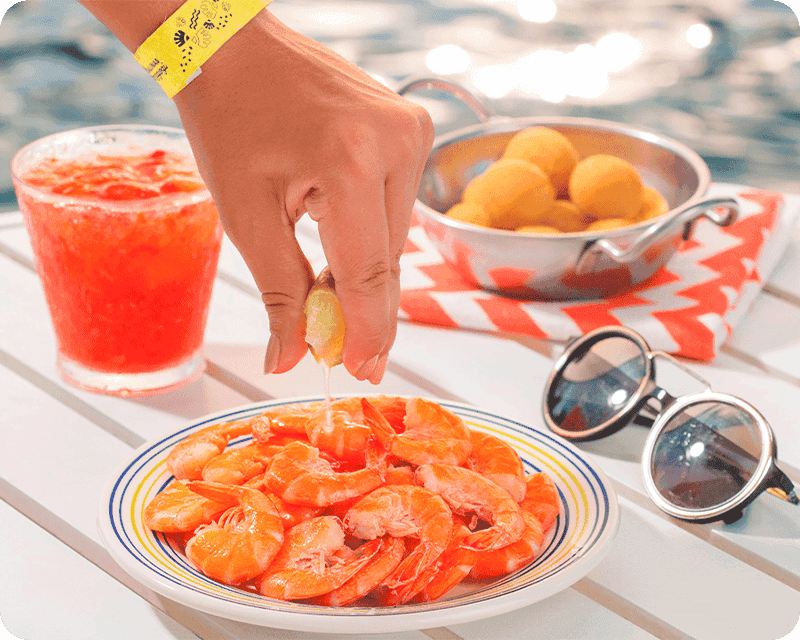 Dois drinks exclusivos do Salinas Maragogi, ao lado de dois pratos de petiscos, um com camarões e o outro com croquetes, sobre mesa à beira da piscina.