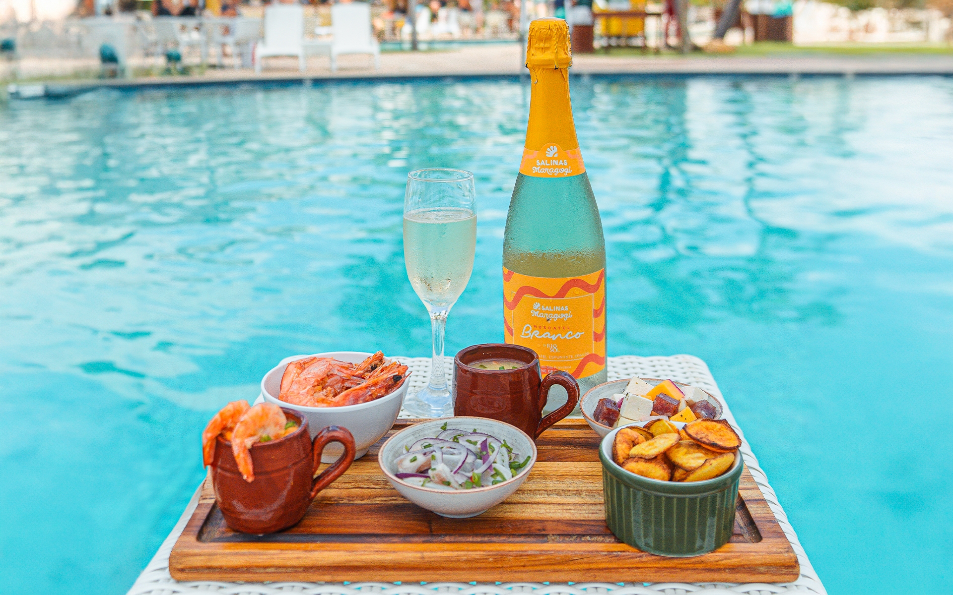 tudo-incluso-na-diária Tábua de madeira com garrafa de Moscatel Branco com uma taça ao lado. Alguns petiscos sendo camarões, ceviche, batatas servidos à beira da piscina. Ao fundo desfocado há a área de lazer, com guarda-sois e espreguiçadeiras.