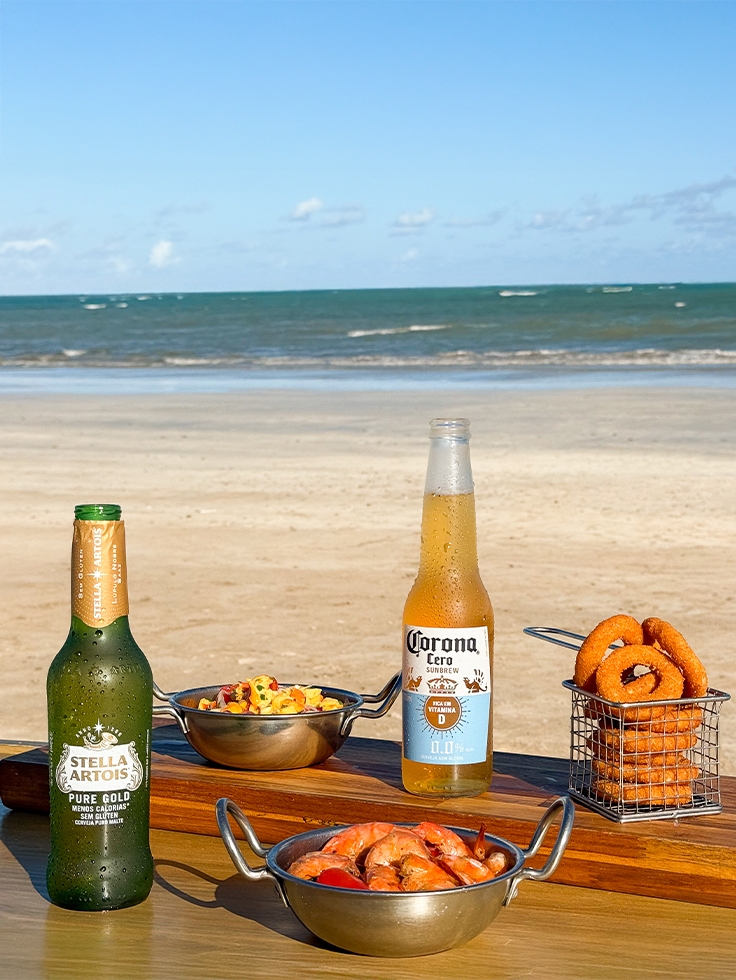 resort-salinas-maragogi-all-inclusive Cervejas Stella Artois e Corona Zero servidas com camarões, anéis de cebola e petiscos em mesa à beira-mar no Salinas Maragogi, com praia e mar ao fundo.