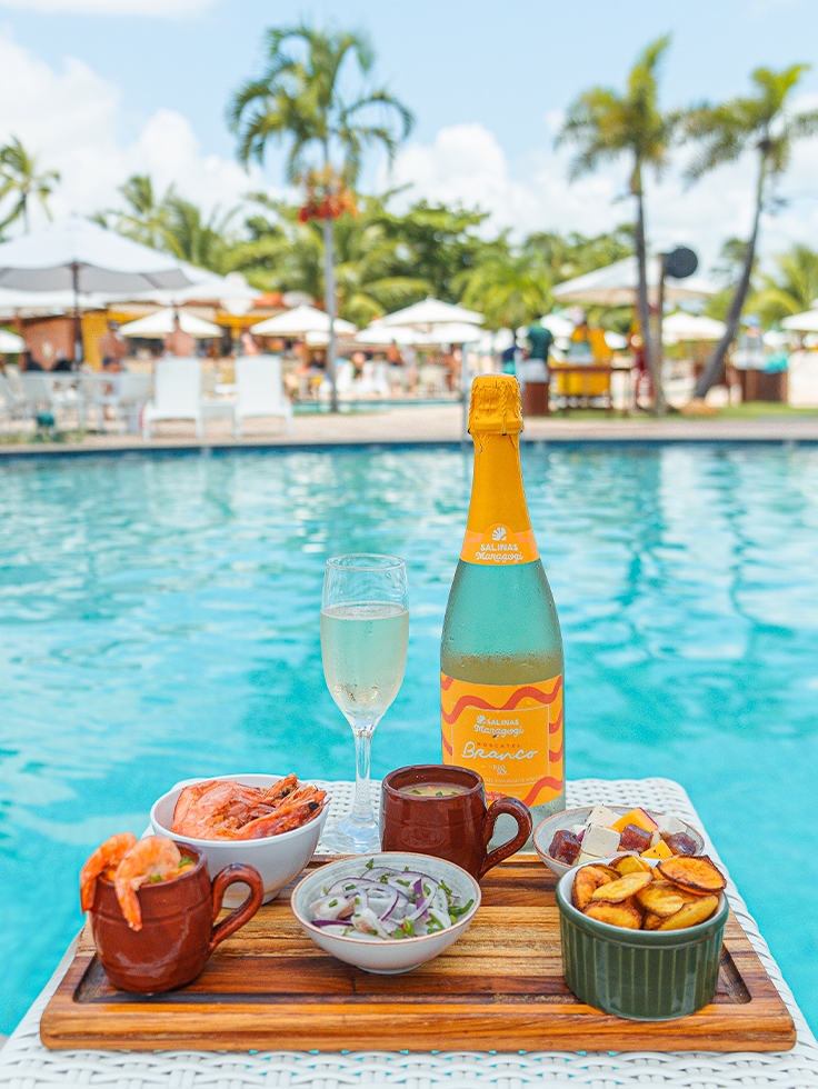 tudo-incluso-na-diária Tábua de madeira com garrafa de Moscatel Branco com uma taça ao lado. Alguns petiscos sendo camarões, ceviche, batatas servidos à beira da piscina. Ao fundo desfocado há a área de lazer, com guarda-sois e espreguiçadeiras.