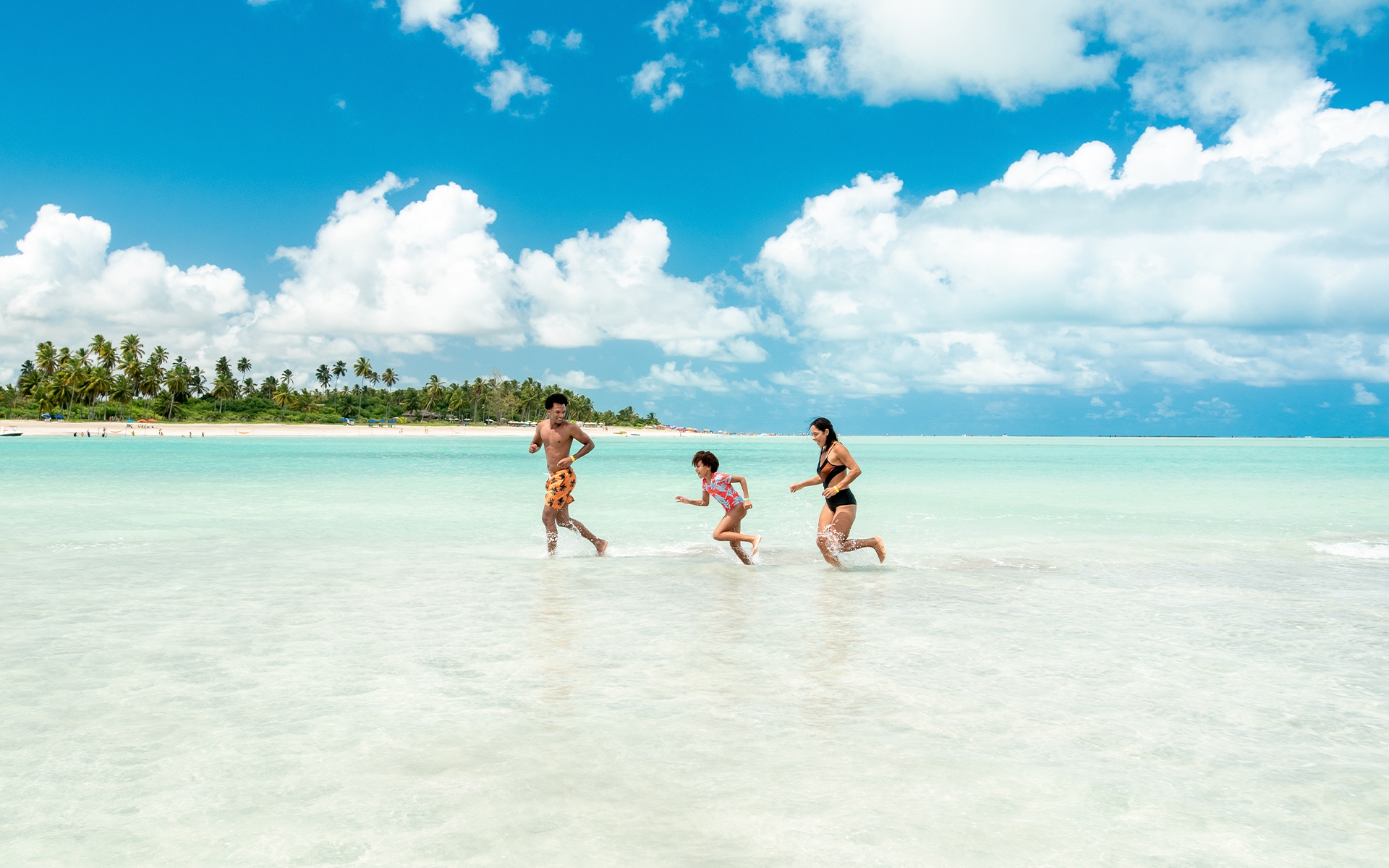praia-de-antunes-maragogi Família correndo em águas rasas e claras da Praia de Antunes, pertinho do Salinas Maragogi. Ao fundo a praia com grandes coqueiros e areia clara.