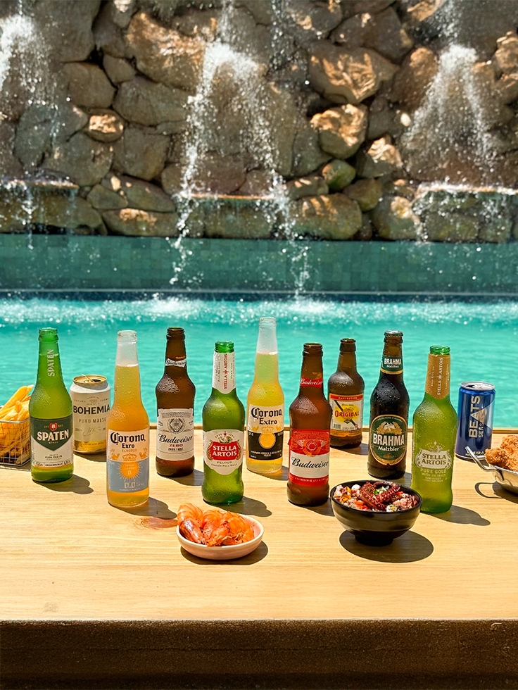 all-inclusive-24h-por-dia Variedade de cervejas long neck oferecidas no resort All Inclusive Salinas Maragogi, sendo Brahma Malzbier, Corona Extra, Beats, Original, Stella Artois, Bohemia Puro Malte e Budweiser, ao lado de diversas tigelas de petiscos, sendo camarões, batatas fritas sobre tábua de madeira à beira da piscina.