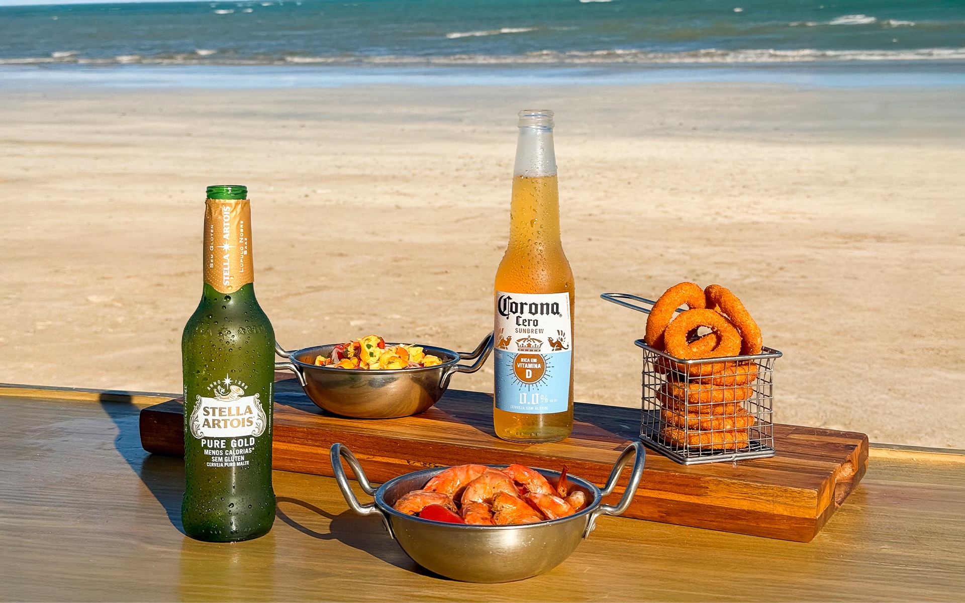 resort-salinas-maragogi-all-inclusive Cervejas Stella Artois e Corona Zero servidas com camarões, anéis de cebola e petiscos em mesa à beira-mar no Salinas Maragogi, com praia e mar ao fundo.