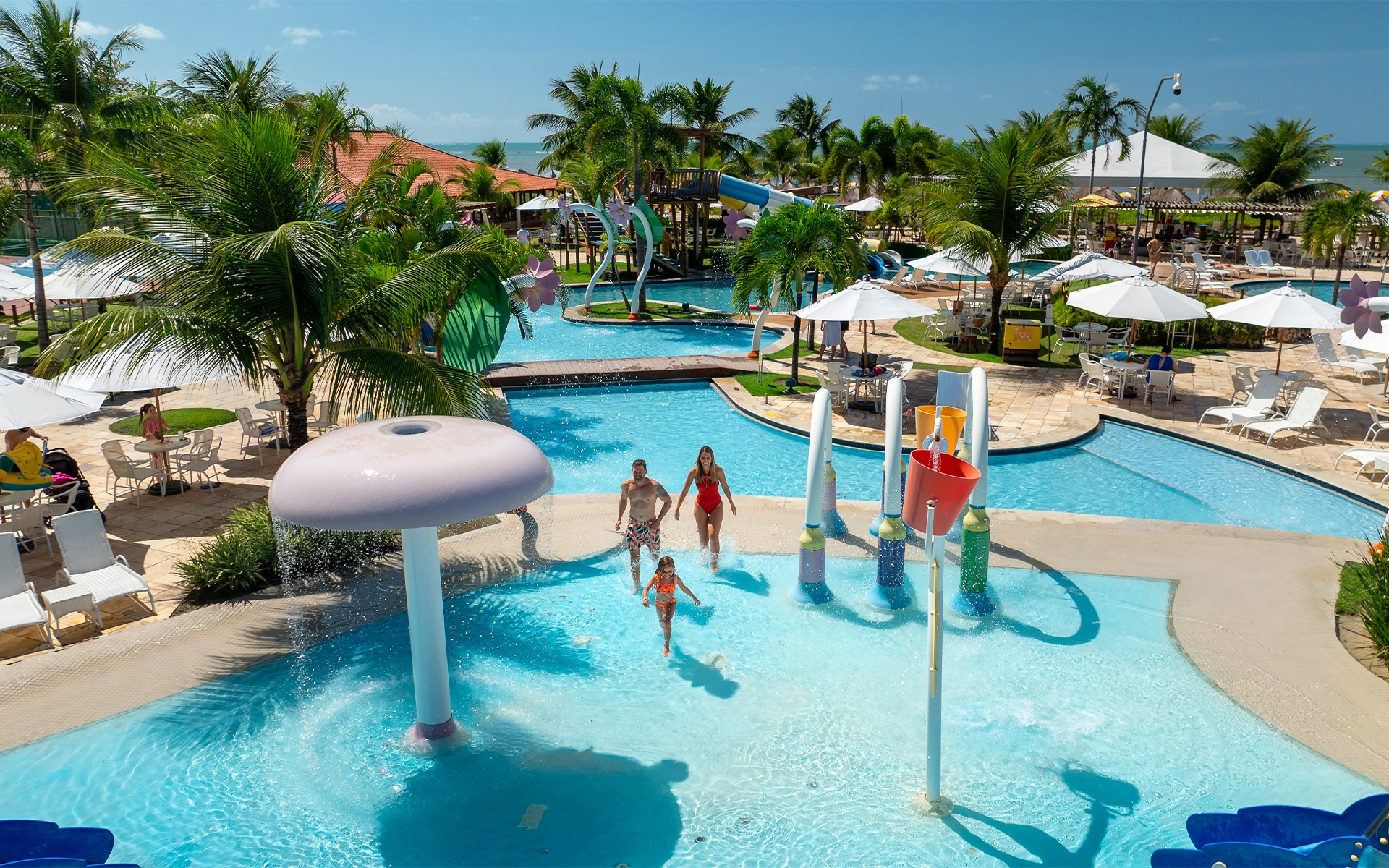 resort-pra-família-na-praia Família aproveitando brinquedos aquáticos da piscina infantil do Salinas Maragogi, com guarda-sóis e espreguiçadeiras ao redor das piscinas em dia de sol.
