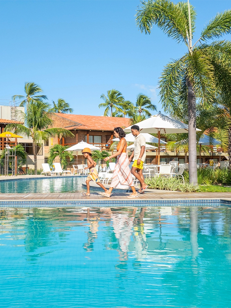 area-de-lazer-salinas-maragogi Família com pai, mãe e criança caminhando pela área de piscinas do Salinas Maragogi, com coqueiros, espreguiçadeiras e guarda-sois ao redor. Ao fundo, um bloco dos apartamentos.