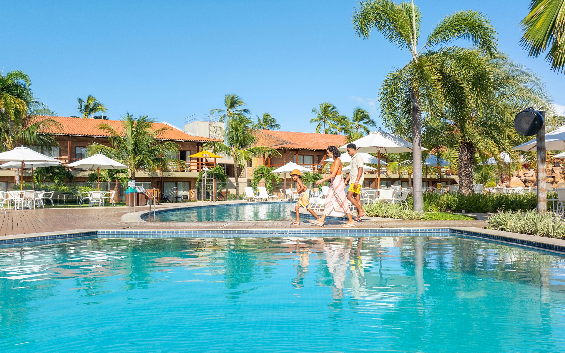 area-de-lazer-salinas-maragogi Família com pai, mãe e criança caminhando pela área de piscinas do Salinas Maragogi, com coqueiros, espreguiçadeiras e guarda-sois ao redor. Ao fundo, um bloco dos apartamentos.