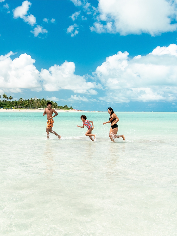 praia-de-antunes-maragogi Família correndo em águas rasas e claras da Praia de Antunes, pertinho do Salinas Maragogi. Ao fundo a praia com grandes coqueiros e areia clara.