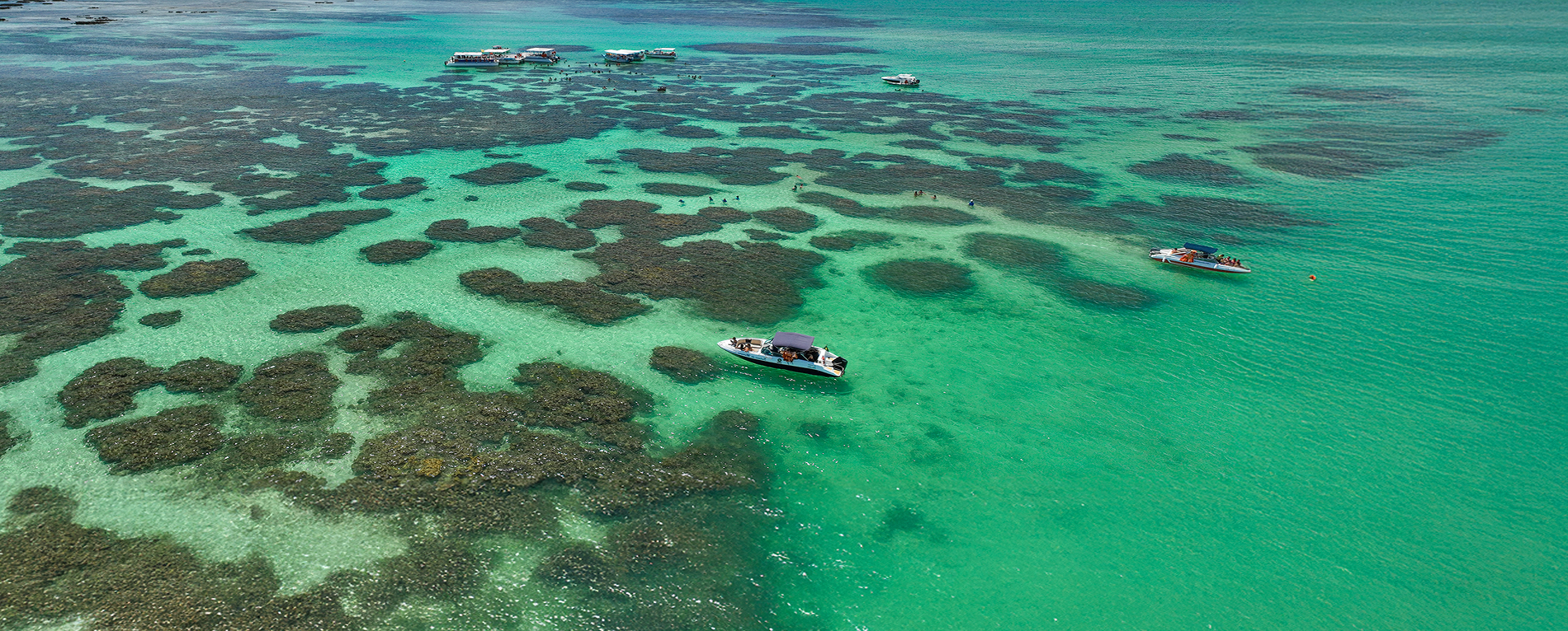 semana-do-consumidor-salinas-maragogi-resort Vista aérea de Galés de Maragogi, com embarcações nas piscinas naturais, tem pessoas nadando na água e é possível ver vários recifes de corais.