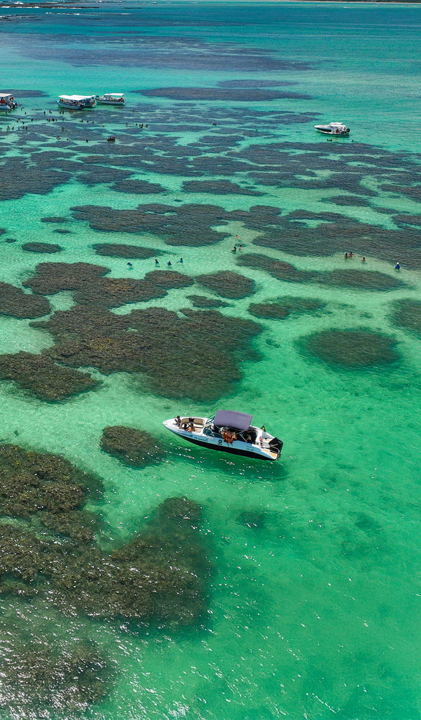 semana-do-consumidor-salinas-maragogi-resort Vista aérea de Galés de Maragogi, com embarcações nas piscinas naturais, tem pessoas nadando na água e é possível ver vários recifes de corais.