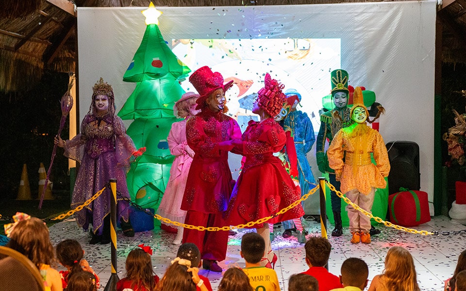 A  foto mostra o final de uma das apresentações das festas natalinas do Salinas Maceió. É possível ver alguns personagens de variadas fantasias e cores como vermelho, roxo, amarelo, rosa e verde. Ao fundo há uma árvore de natal e há crianças atentas assistindo a apresentação.
