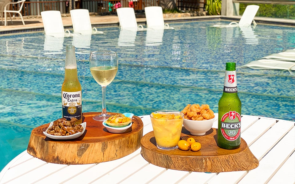 Na foto há duas bandejas de madeira na beira de uma das piscinas do resort Salinas Maceió. Há petiscos e bebidas que fazem parte do all inclusive: uma long neck corona, petiscos de carne, camarões fritos e quiche. Há também uma caipirinha e uma long neck becks.