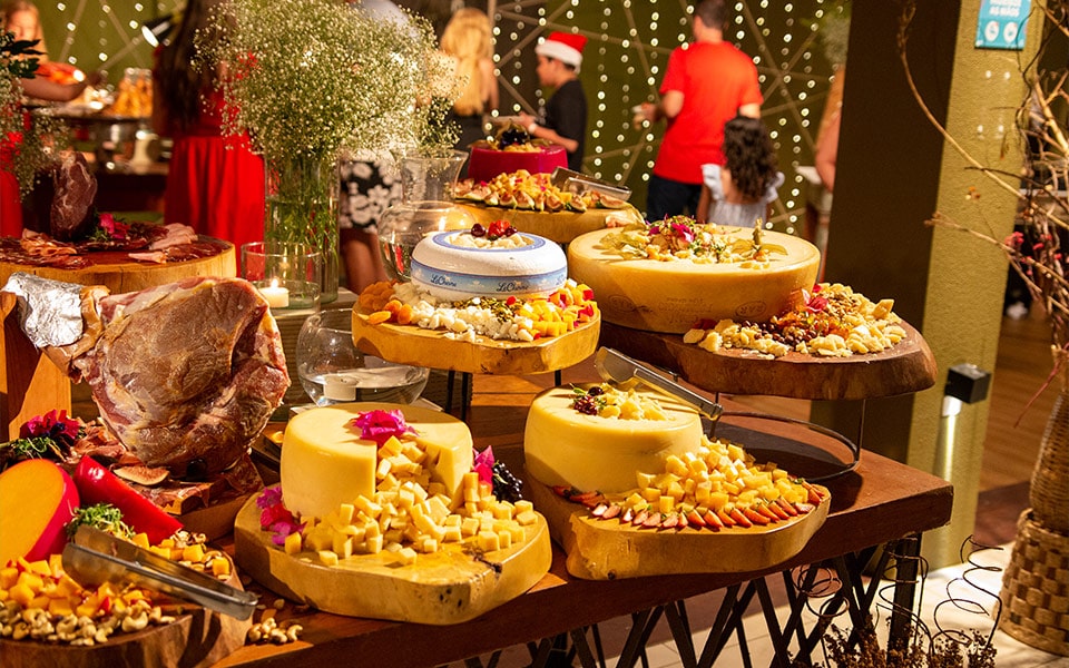 Uma mesa de frios repleta de queijos fazendo parte da ceia especial do natal do Salinas Maceió. É possível ver várias peças de queijos decorados com acessórios natalinos, assim como embutidos e uma peça de presunto do tipo parma. Há também um vaso de florzinhas em cima da mesa.