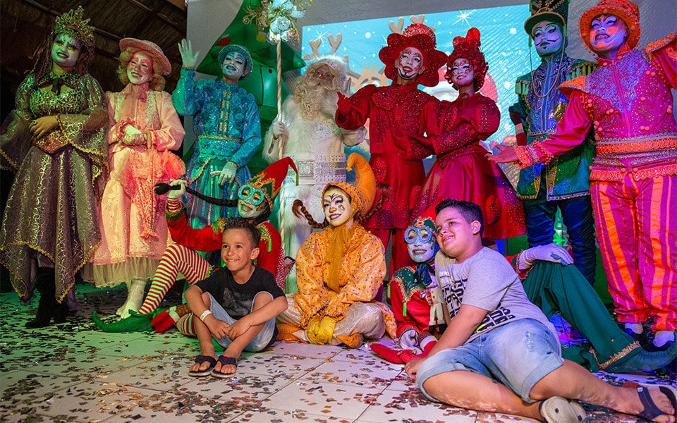 Na foto duas crianças pousam sorridentes para uma foto com os personagens da apresentação especial de natal do Salinas Maceió. Há diversos personagens de cores variadas como verde, rosa, azul, laranja e ver. O papai noel está no centro com barba e roupa totalmente branca.