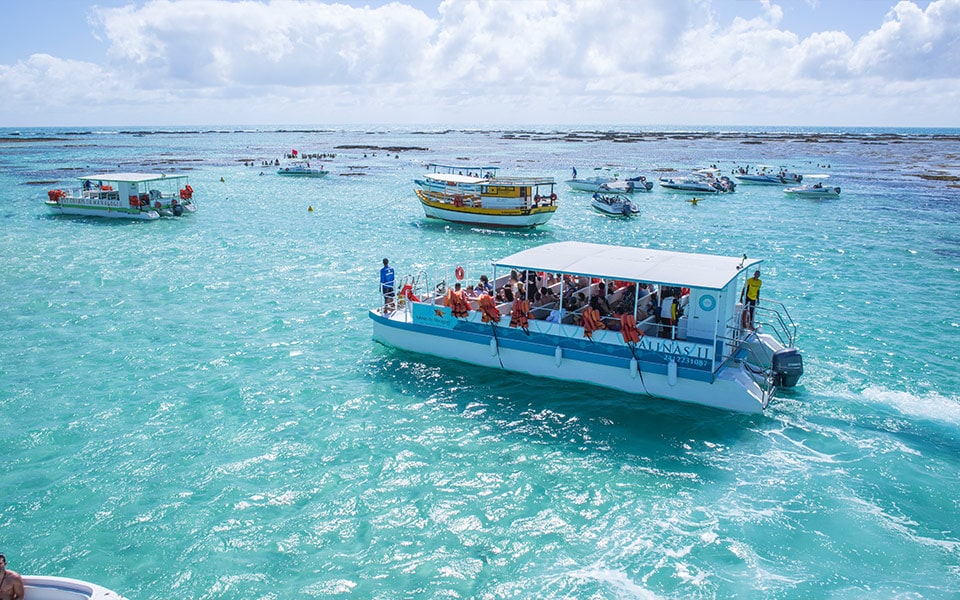 Na foto é possível ver a Galés de Maragogi com suas águas azuis e cristalinas. Há muitos catamarãs e em evidência um grande catamarã com vários turistas que estão a passeio pelo Salinas.