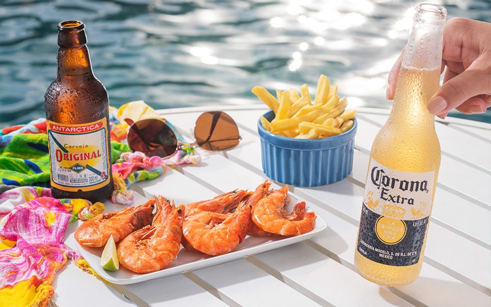 Na foto é possível ver petiscos e bebidas na beira da piscina de Salinas Maragogi. Há camarões ao alho e óleo, uma porção de batata frita, uma cerveja long neck Corona, uma cerveja long neck antarctica original, uma canga estampada e um óculos de sol.