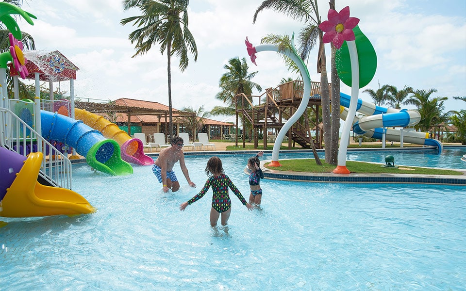 Na foto é possível ver um pai que brinca com as duas suas filhas em uma fas piscinas do salinas maragogi. É possível ver escorregadores ao fundo que garantem muita diversão em família.