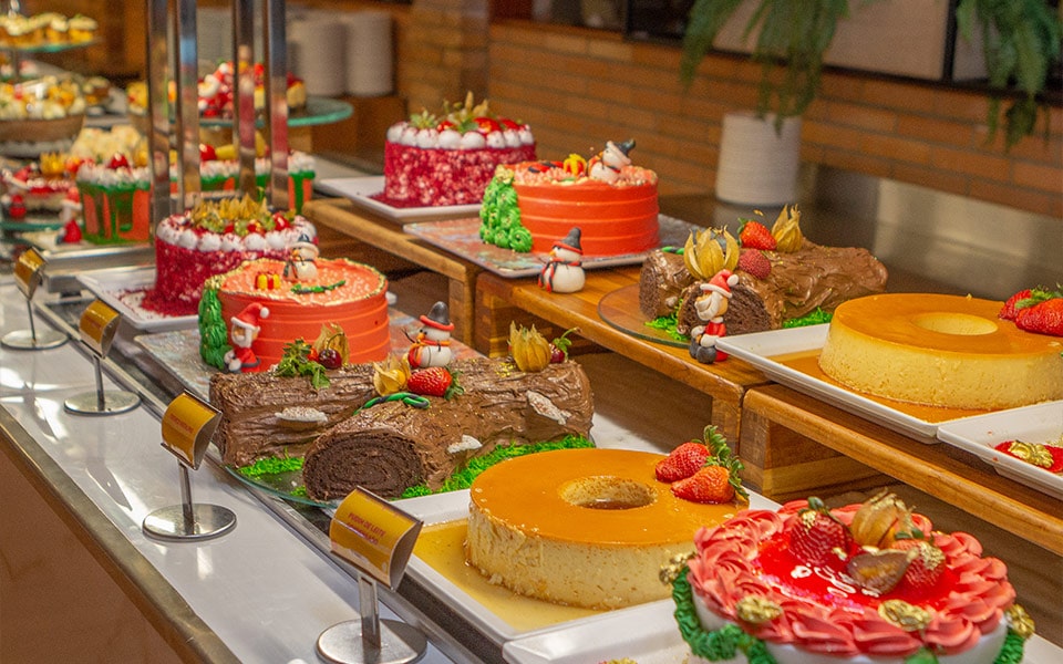 Na foto é possível ver uma mesa de sobremesas do restaurante principal do salinas maragogi. Há cerca de 9 tortas diferentes e temáticas. Entre elas é possível ver clássicos brasileiros como pudim, rocambole de chocolate, tortas de morango e red velvet, todas decoradas com 